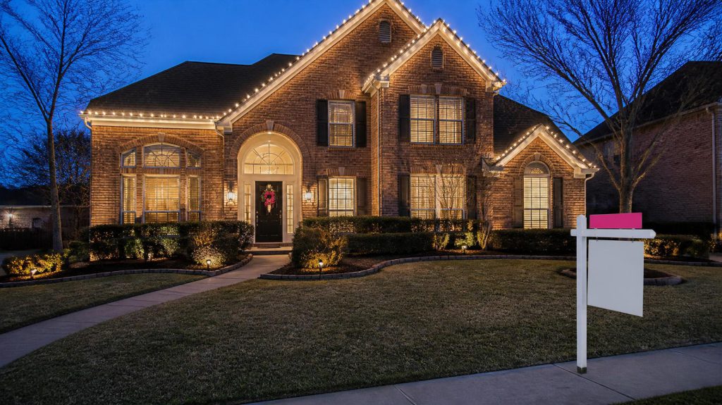 Two-story brick Katy home at dusk with white holiday lights, a pink-bowed wreath on the door, and a for sale sign with a hot pink rider in the front yard.